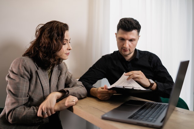 landlord-with-curly-brown-hair-in-a-brown-plaid-suit-reviews-a-rental-agreement-with-their-new-tenant-who-holds-a-clipboard-with-a-document-o- it