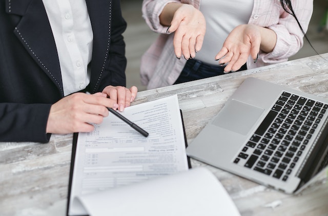 A-Person-Holding-Black-Pen-pointing-something-on-a-paper-discussing-with-someone-else-in-front-of-a-computer